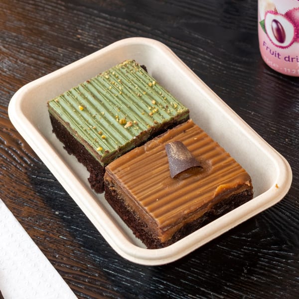 Two brownies, one matcha and one caramel, in a light brown tray on a dark wooden table.