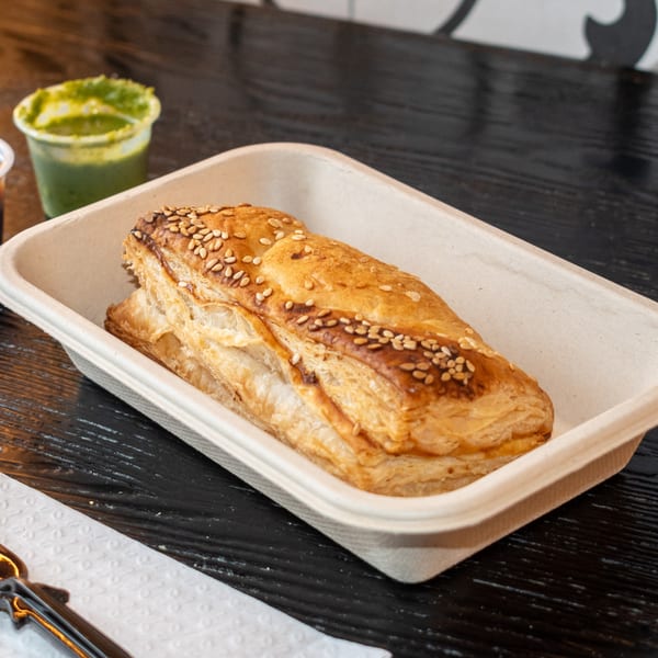 Golden brown Bombay Baking Co. puff pastry with sesame seeds in a container, with green chutney.
