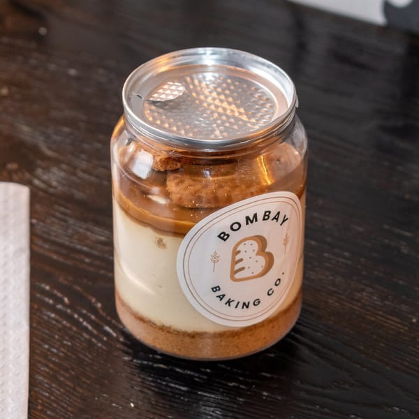 A Bombay Baking Co. dessert jar with layers of cream, crumbs, and caramel on a dark wooden table.