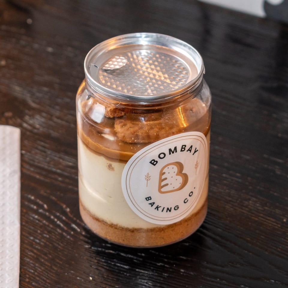 A Bombay Baking Co. dessert jar with layers of cream, crumbs, and caramel on a dark wooden table.