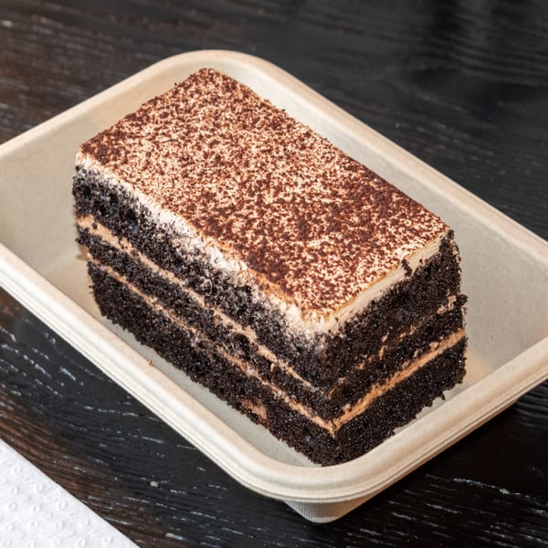 A rectangular slice of chocolate cake with layers of frosting and cocoa powder on top, in a paper tray.