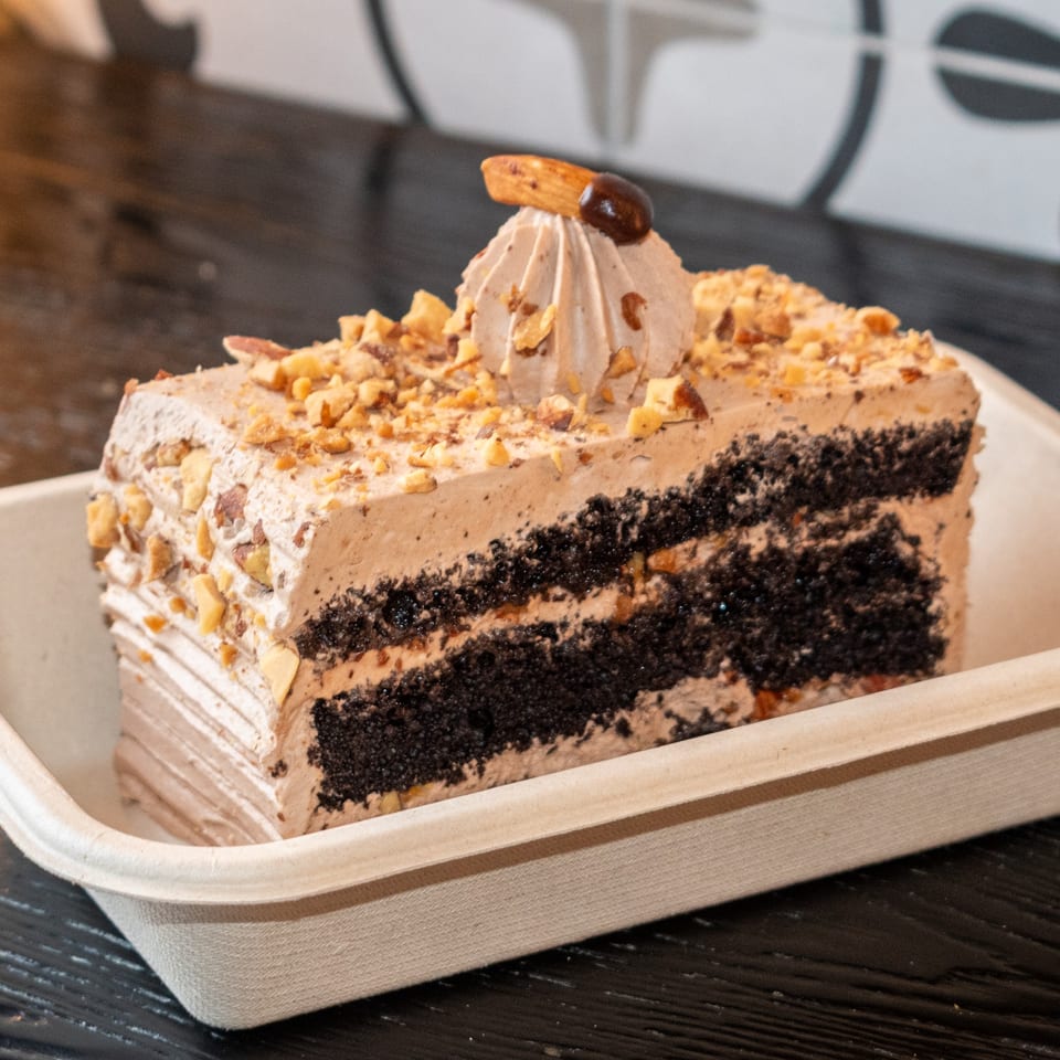 A slice of chocolate cake with light brown frosting, topped with nuts and a decorative swirl.