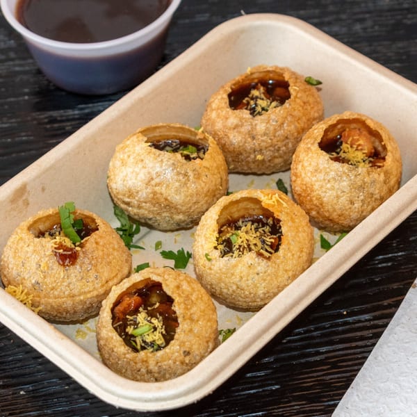 Six pani puri, filled with dark sauce and toppings, in a light brown tray with a sauce cup.