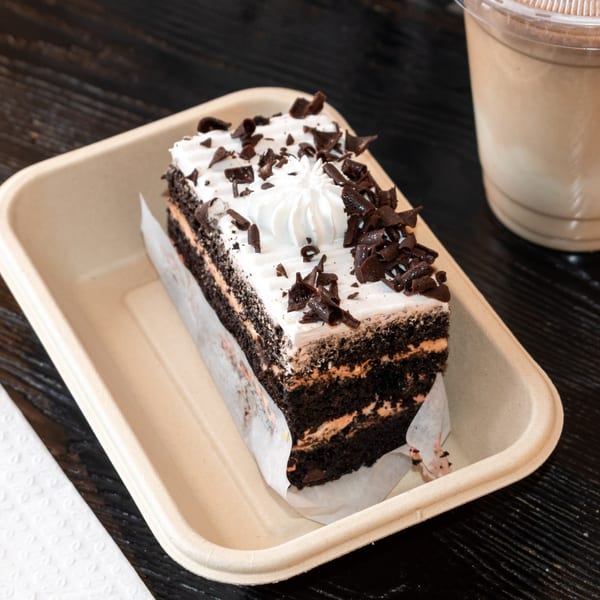 A slice of chocolate cake with white frosting and chocolate shavings in a to-go container.