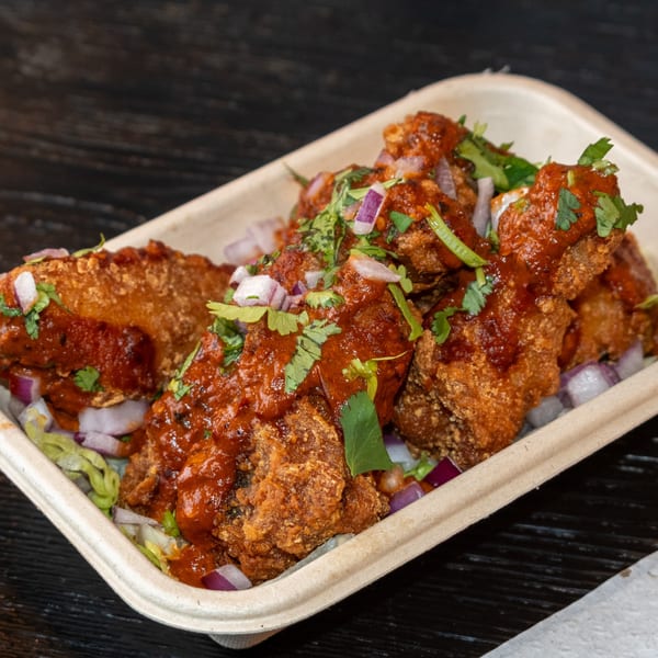 Crispy fried chicken pieces topped with red sauce, cilantro, and diced red onions in a container.