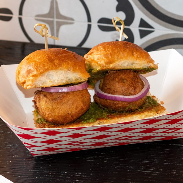 Two Bombay Baking Co. vada pav sliders with red onion and green chutney in a paper tray.