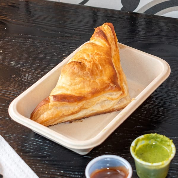 A golden-brown, flaky pastry from Bombay Baking Co. in a light brown tray with two dipping sauces.