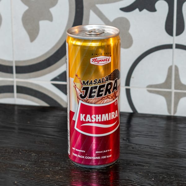 A Hajuri Masala Jeera Kashmira soda can, gold and red, on a dark wooden surface.