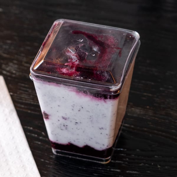 A clear plastic container filled with white yogurt and dark berry compote, on a dark wooden table.