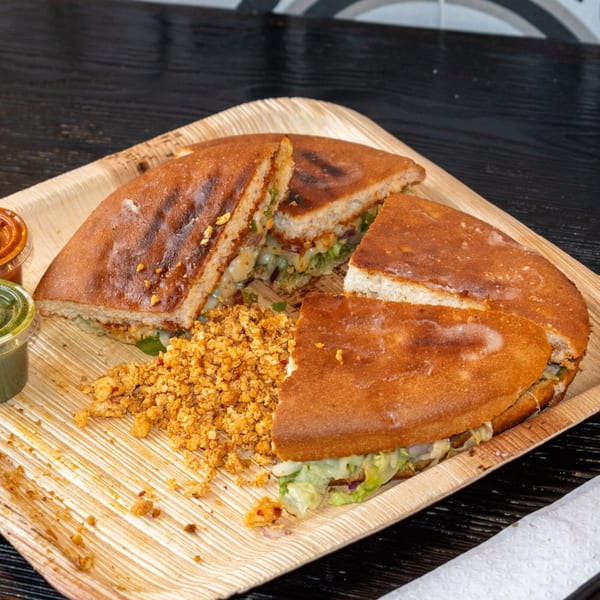 A round, grilled sandwich cut into quarters, with a side of crumbled paneer and two sauces.