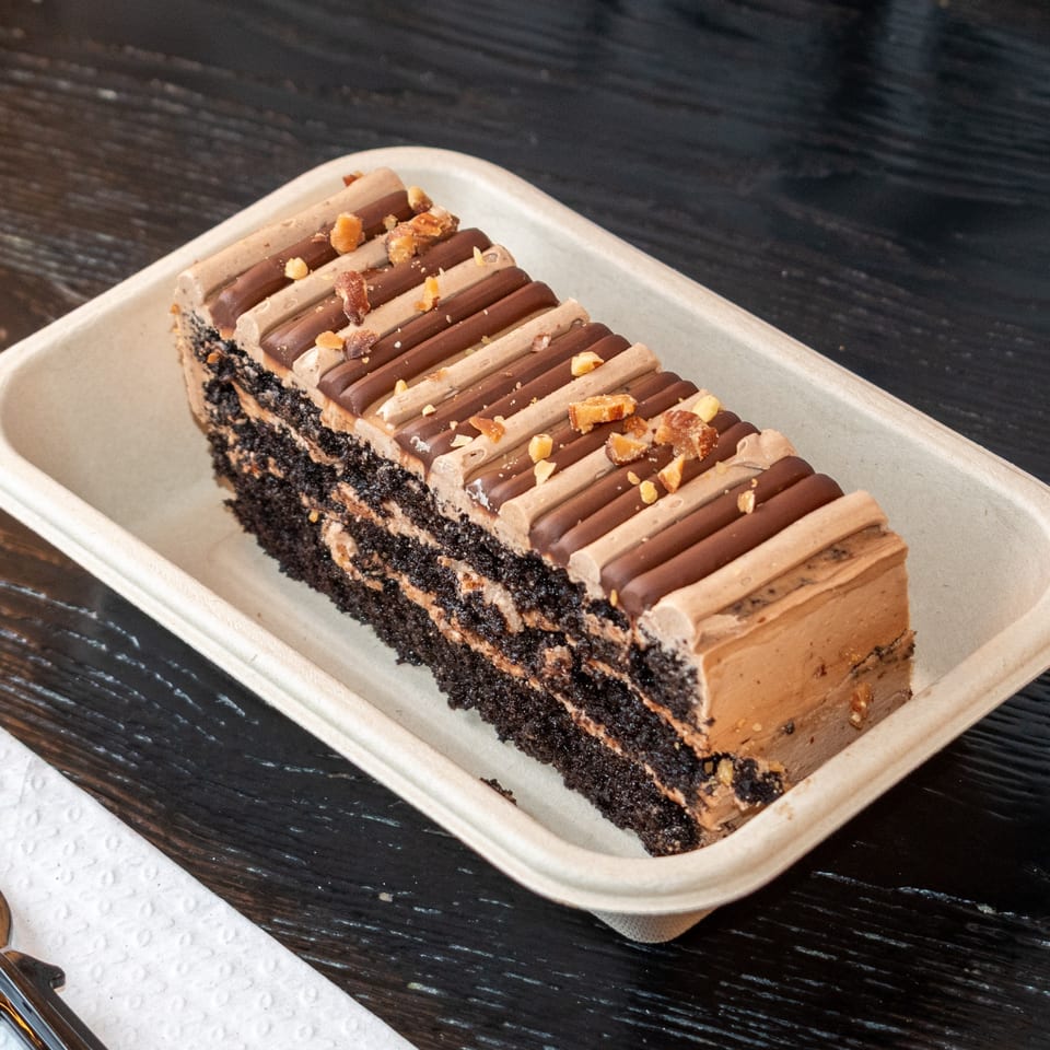 A rectangular slice of chocolate cake with layers of frosting and chocolate bars, topped with nuts.