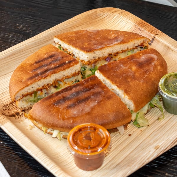 Bombay Baking Co.'s grilled sandwich, cut into four pieces, served with two dipping sauces on a wooden tray.