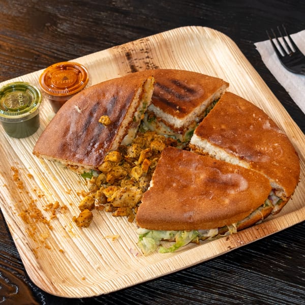 A round, grilled chicken sandwich, cut into quarters, with two sauces on a wooden plate.