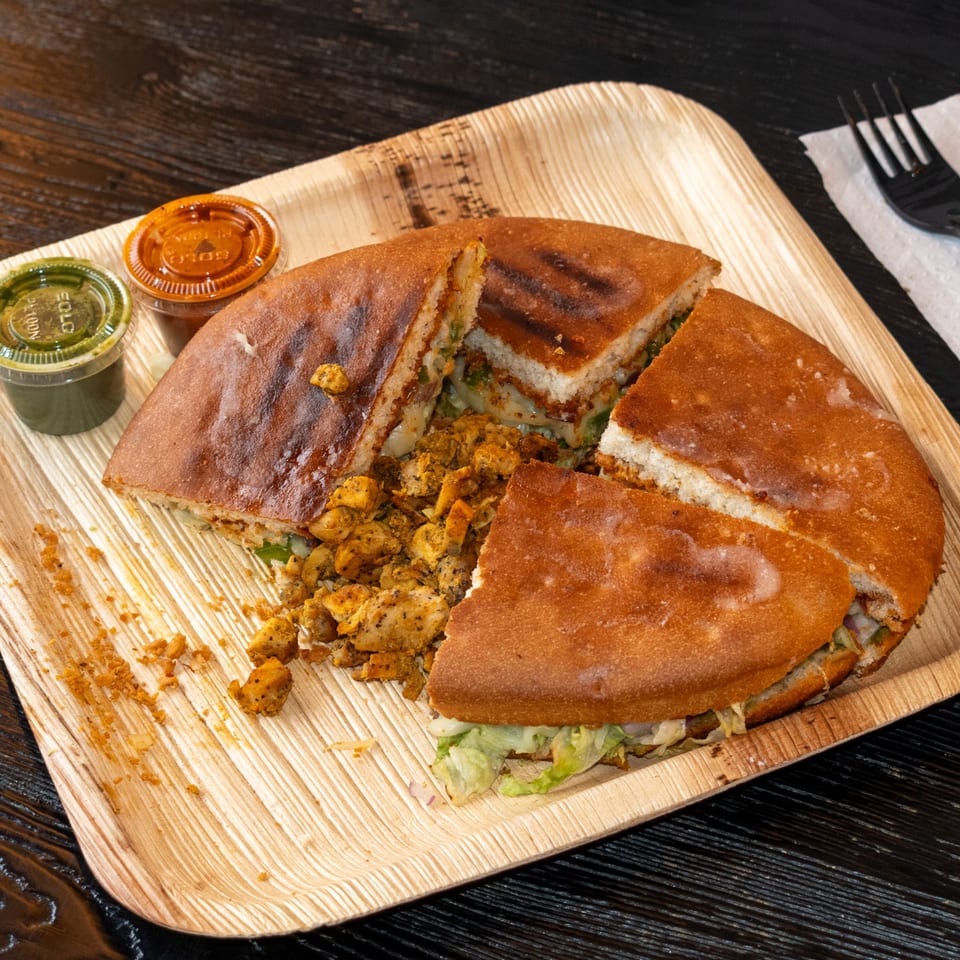 A round, grilled chicken sandwich, cut into quarters, with two sauces on a wooden plate.