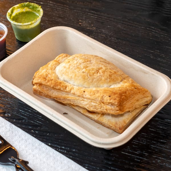 Golden-brown Bombay Baking Co. puff pastry in a tray with green chutney on a dark wooden table.