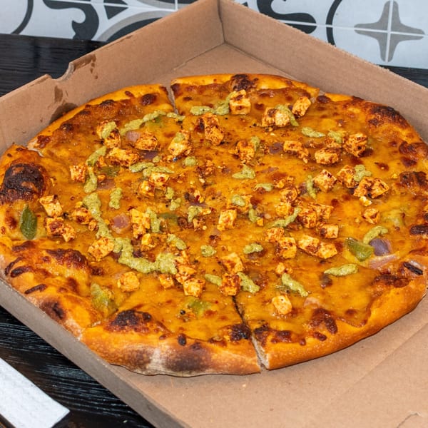 A Bombay Baking Co. paneer tikka pizza with green chutney drizzle in a cardboard box.