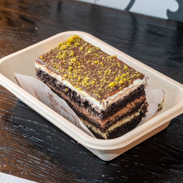 A rectangular chocolate cake with pistachio sprinkles sits in a beige takeout container.