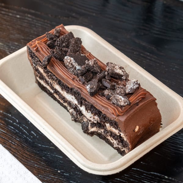 A rectangular slice of chocolate cake with cream filling and cookie crumbles in a beige container.