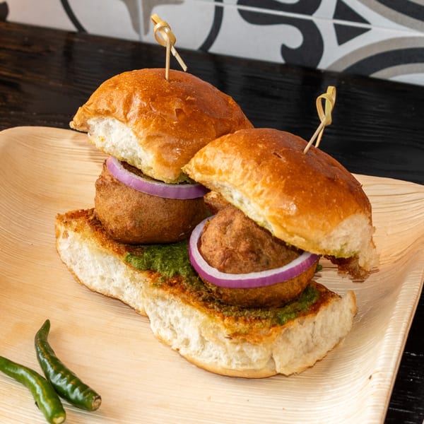 Two Bombay Baking Co. vada pav sliders with green chutney and red onion on a wooden plate.