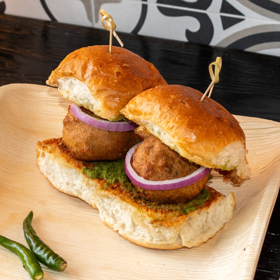 Two Bombay Baking Co. vada pav sliders with green chutney and red onion on a wooden plate.