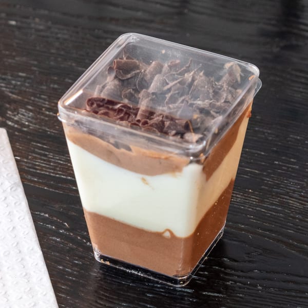 Layered chocolate and vanilla dessert in a clear plastic cup, topped with chocolate shavings.