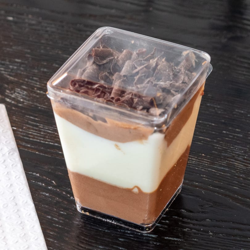 Layered chocolate and vanilla dessert in a clear plastic cup, topped with chocolate shavings.