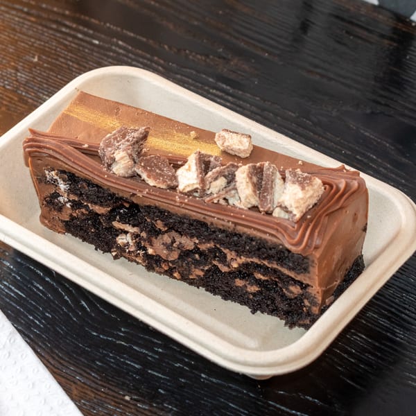 A slice of chocolate cake with chocolate frosting and candy pieces in a beige container.