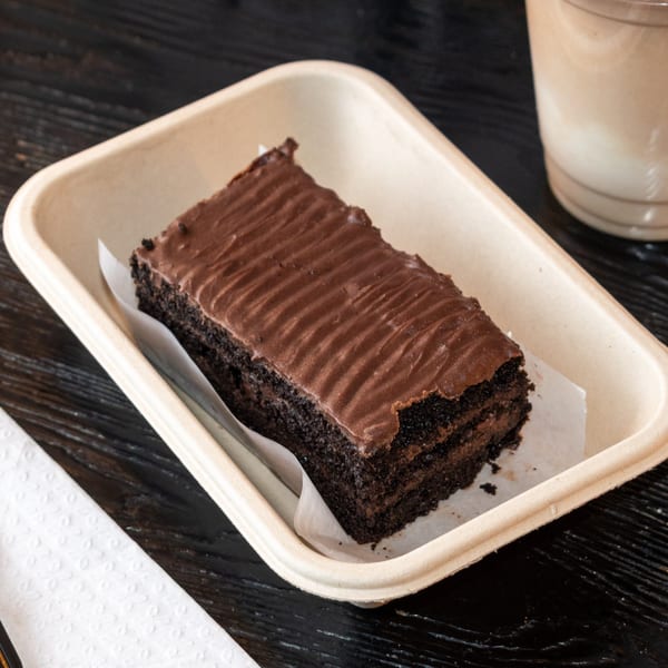 A slice of rich chocolate cake with frosting in a light brown tray on a dark wooden table.