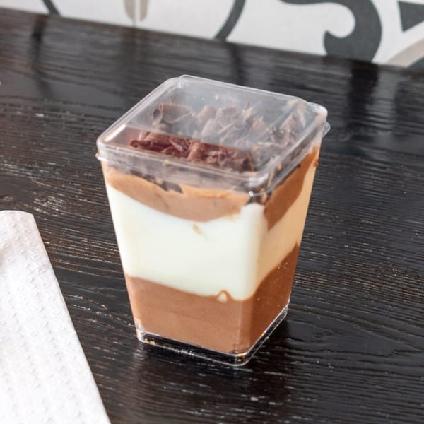 Layered chocolate and vanilla dessert in a clear plastic cup with chocolate shavings on top.