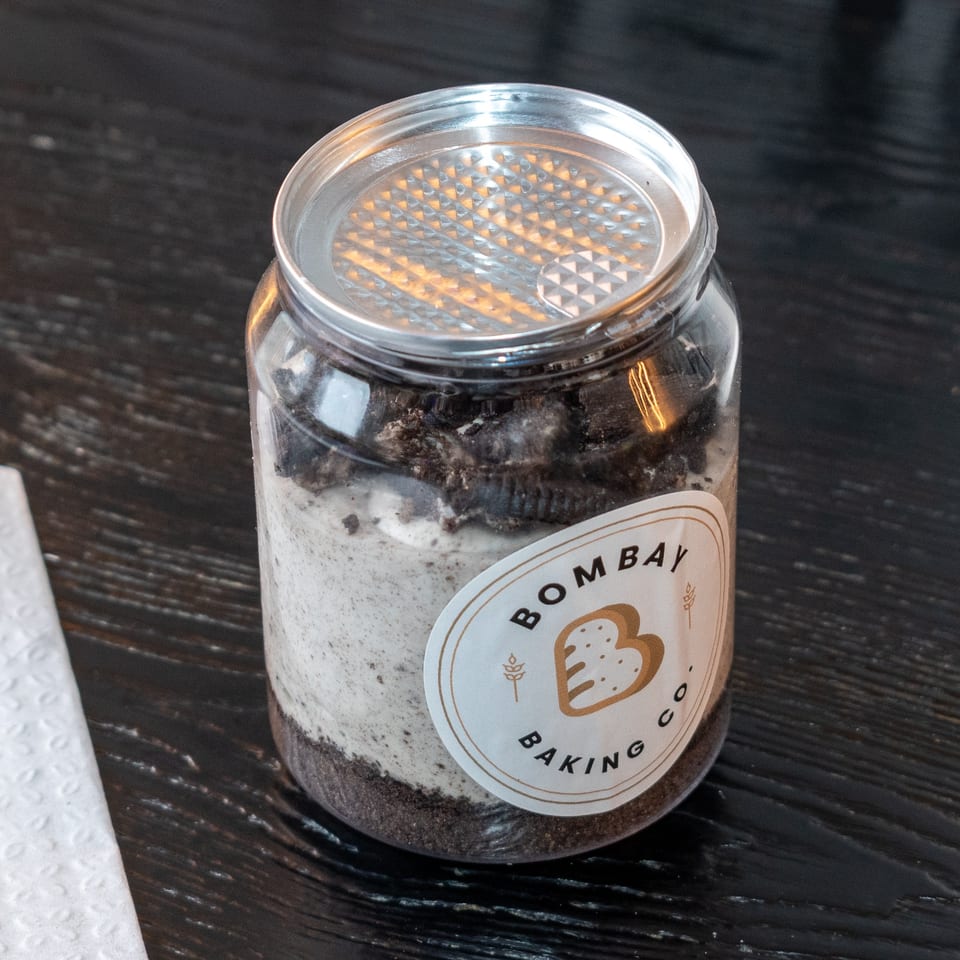 Bombay Baking Co. dessert jar with layers of cream and cookie crumbles on a dark wooden table.