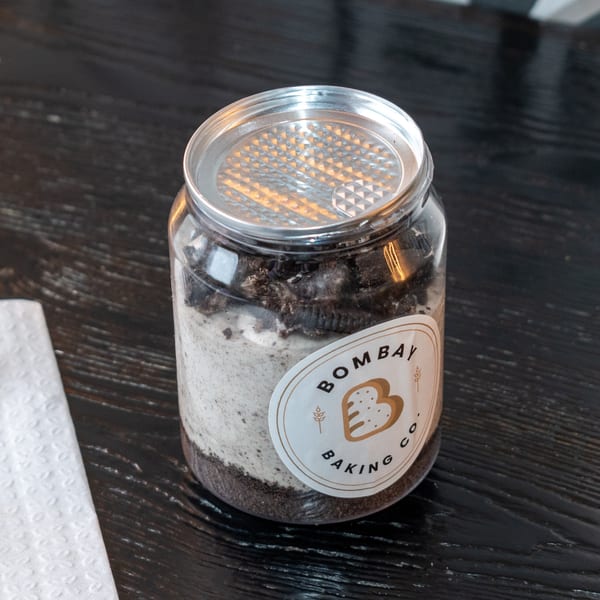 A Bombay Baking Co. dessert jar with layers of cream and cookie crumbles on a dark wooden table.