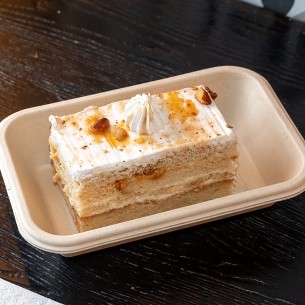A slice of Bombay Baking Co. cake with white frosting, caramel drizzle, and nuts in a to-go container.
