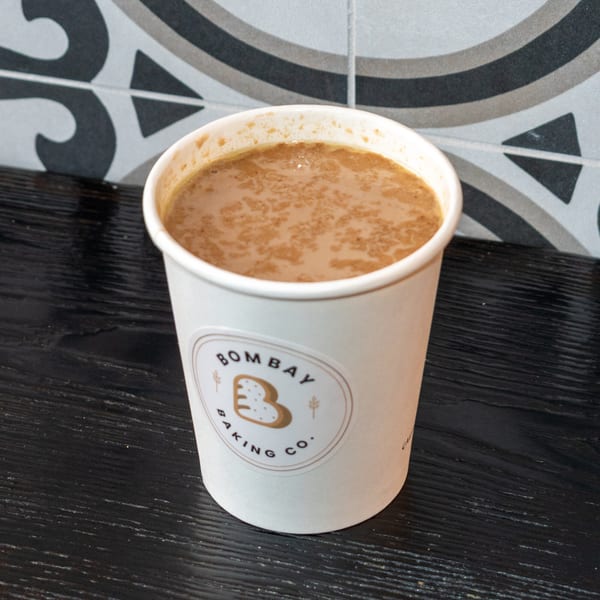 A Bombay Baking Co. branded cup filled with a light brown beverage, possibly chai, on a dark surface.