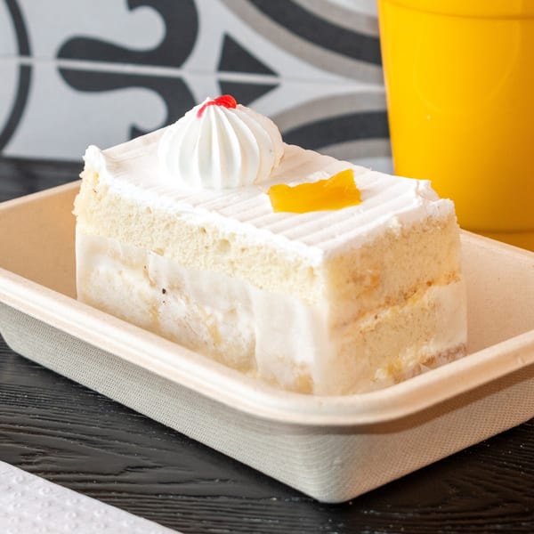 A slice of white cake with whipped cream, a red cherry, and a mango piece in a to-go container.