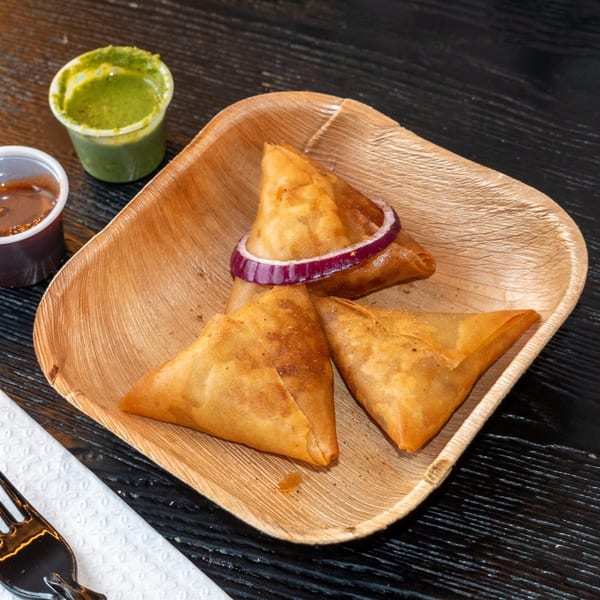 Three golden-brown samosas with a red onion ring on a wooden plate, with two dipping sauces.