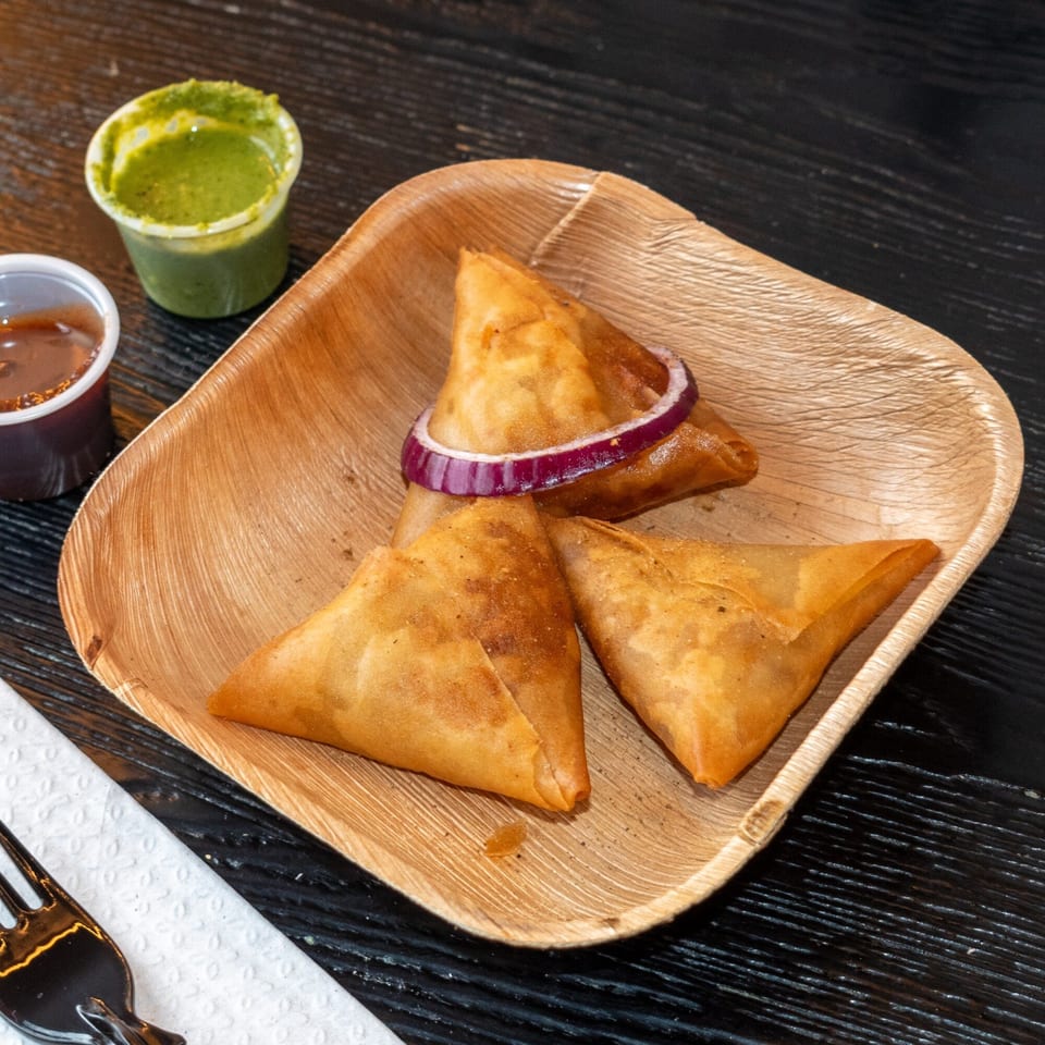 Three golden-brown samosas with a red onion ring on a wooden plate, with two dipping sauces.