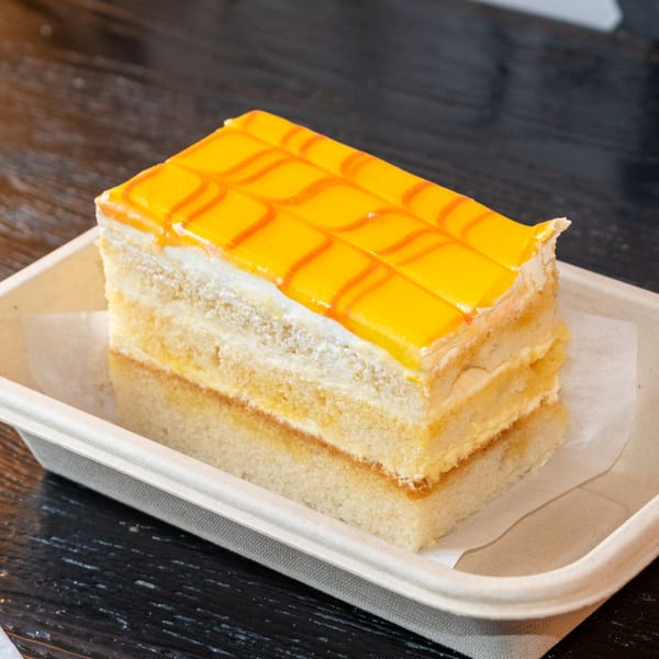 A rectangular slice of Bombay Baking Co.'s mango cake with yellow icing and orange drizzle.