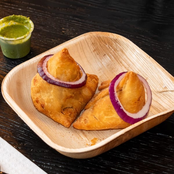 Two golden-brown samosas topped with red onion slices, served on a wooden plate with green chutney.