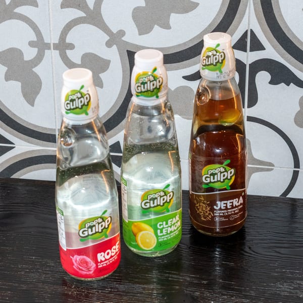 Three Pop & Gulpp soda bottles: Rose, Clear Lemon, and Jeera, on a dark surface.