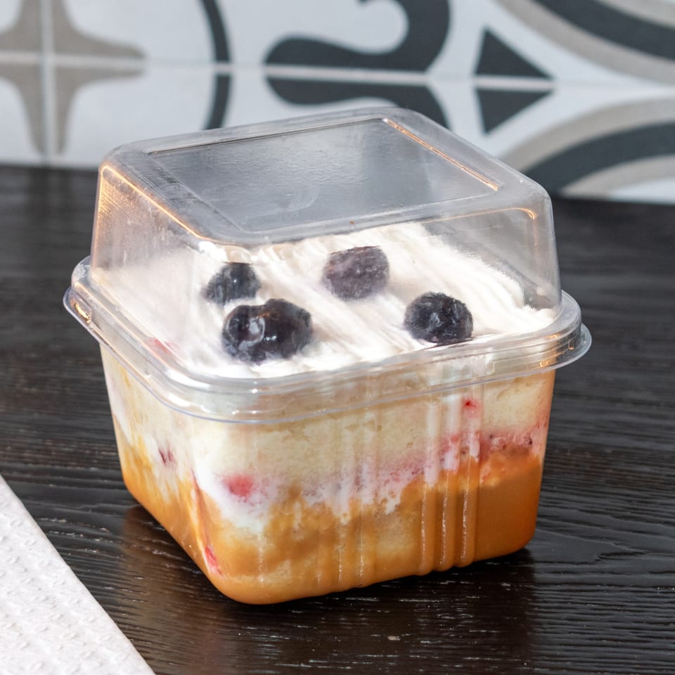A clear plastic container holds a layered dessert with cake, fruit, cream, and blueberries.