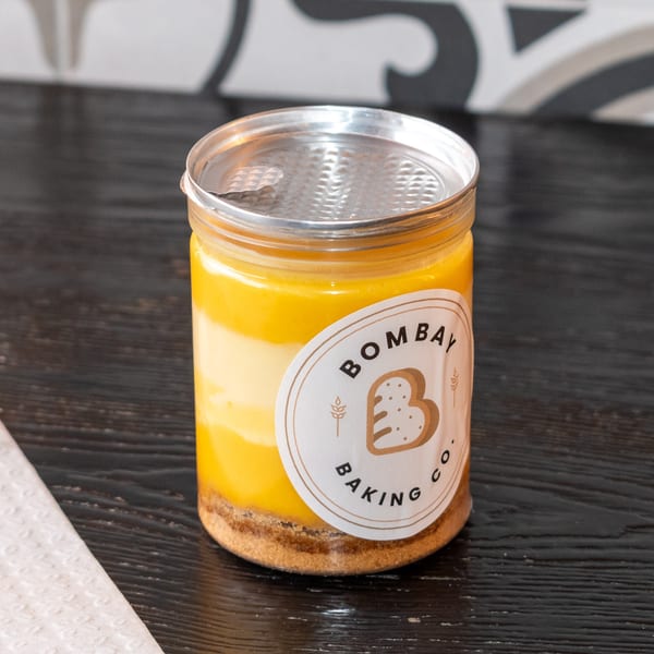 A Bombay Baking Co. mango cheesecake in a clear jar with a silver lid on a dark wooden surface.