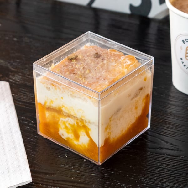 A square plastic container holds a layered dessert with white and orange sections, topped with nuts.