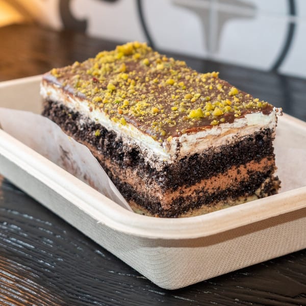 A rectangular slice of chocolate cake with white frosting, chocolate ganache, and pistachio crumbles.