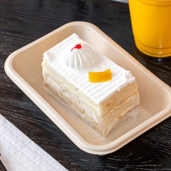 A slice of Bombay Baking Co.'s vanilla cake with white frosting, a red cherry, and yellow garnish.