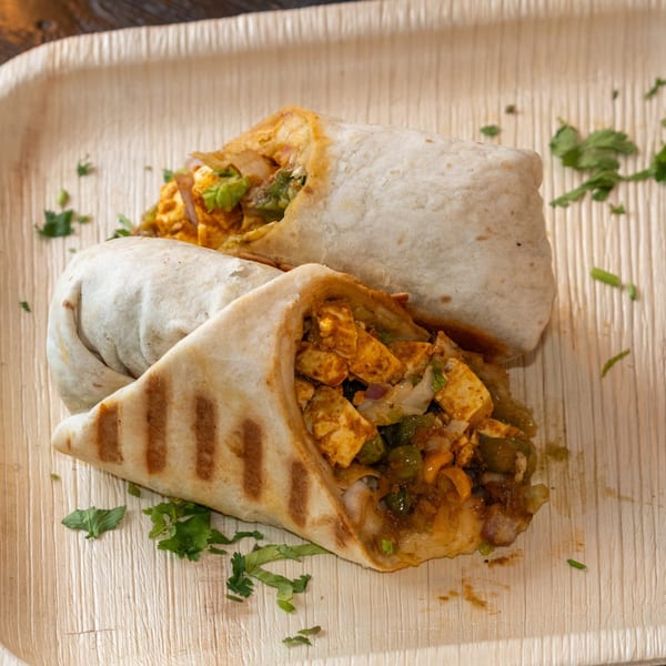 Two Bombay Baking Co. paneer wraps, one grilled, on a light wooden plate with cilantro.