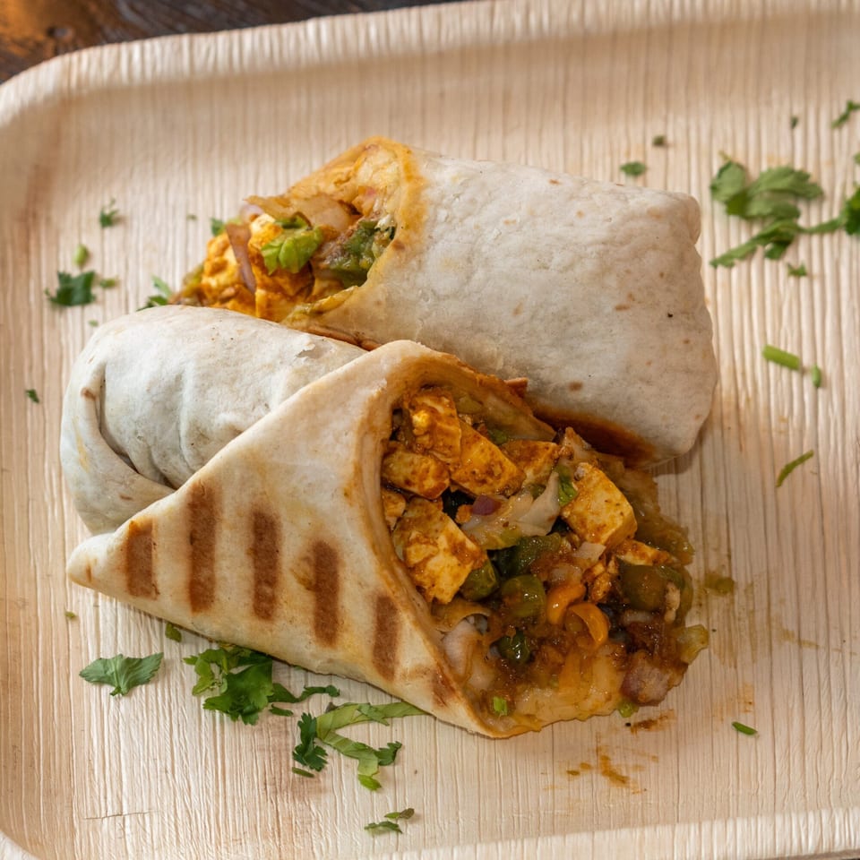Two Bombay Baking Co. paneer wraps, one grilled, on a light wooden plate with cilantro.