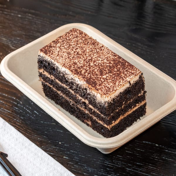 A rectangular slice of chocolate cake with frosting and cocoa powder on top, in a beige container.