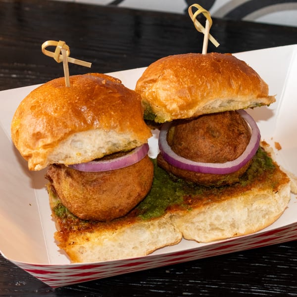 Two vada pav sliders with red onion and green chutney in a white paper tray.