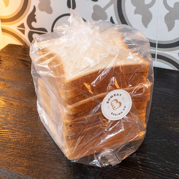 A plastic-wrapped loaf of Bombay Baking Co. sliced bread on a dark wooden surface.