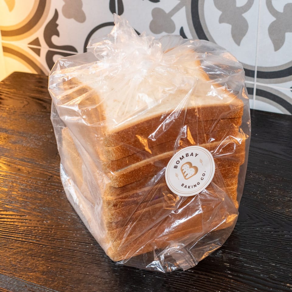 A plastic-wrapped loaf of Bombay Baking Co. sliced bread on a dark wooden surface.